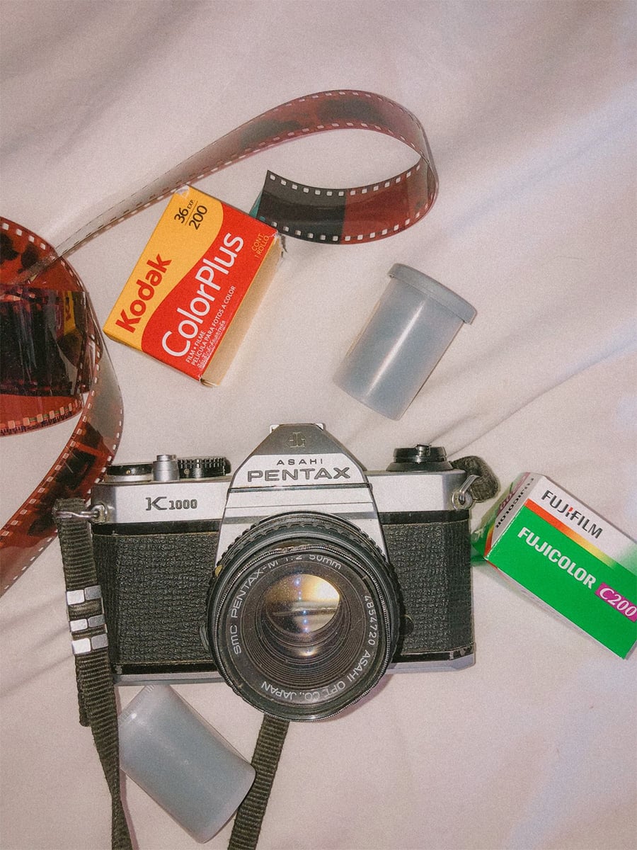 A Pentax film camera with exposed film, Kodak ColorPlus and Fujifilm FujiColor film boxes, and two empty film canisters on a white surface.