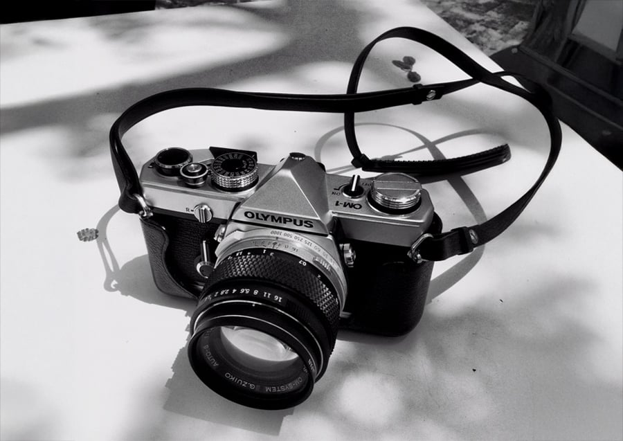 A black and white photo of an Olympus OM-1 film camera with a strap, resting on a white surface, with shadows of leaves in the background.