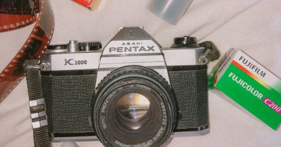 A Pentax K1000 film camera sits on a light surface, surrounded by a roll of film, a film canister, and a box of Fujicolor C200 film.