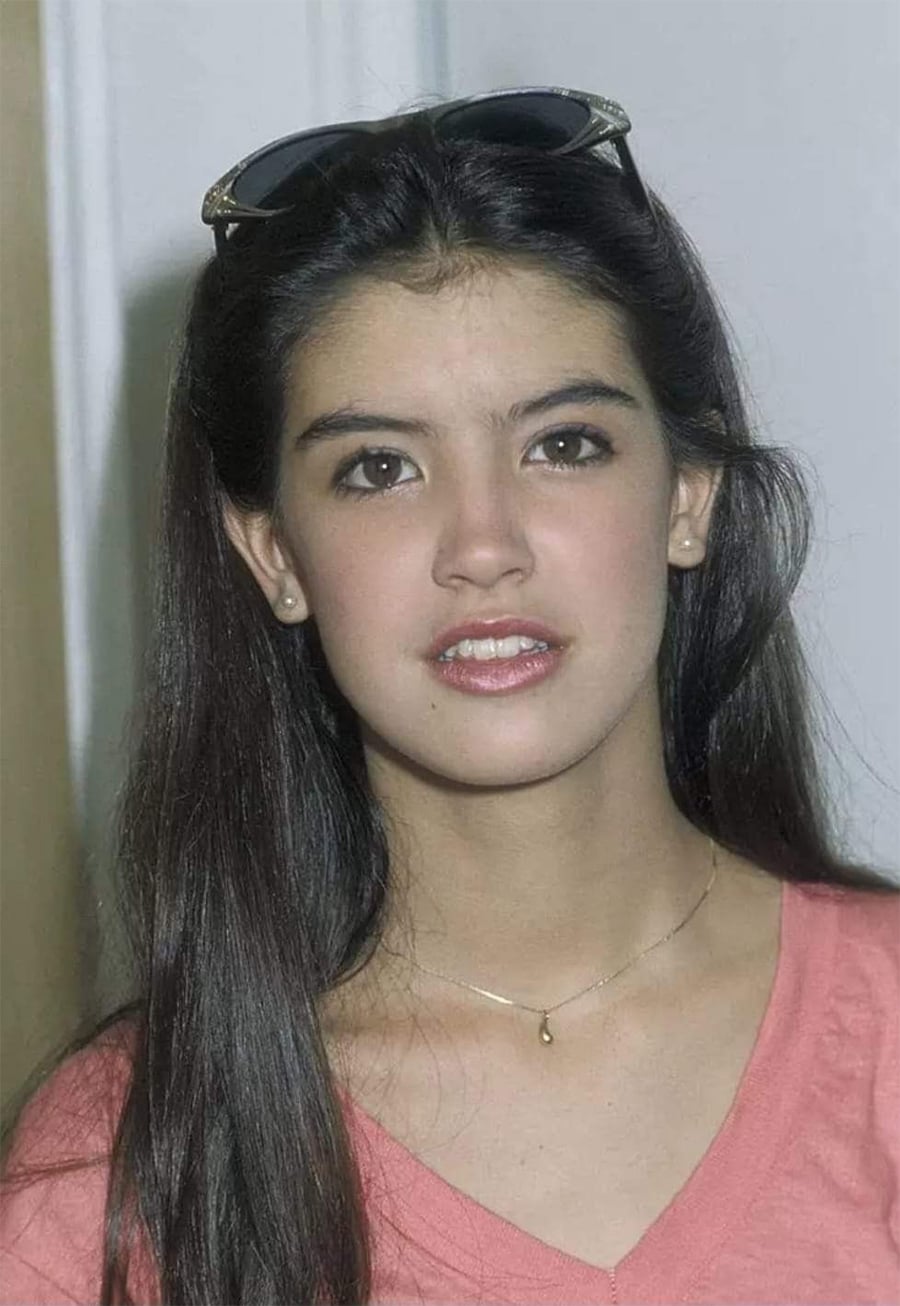 A young woman with long dark hair, wearing sunglasses on her head, a pink top, and a delicate necklace, stands indoors and looks at the camera with a neutral expression.