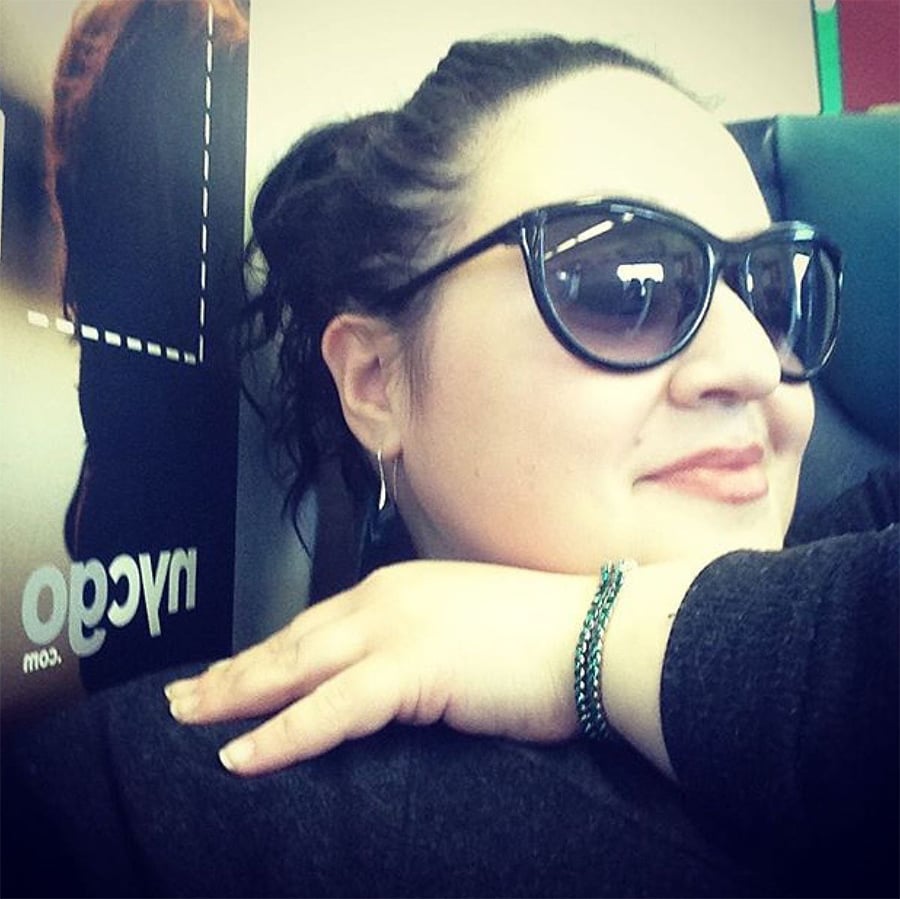 Woman with dark hair and sunglasses leans her head on her hand, wearing a bracelet and smiling slightly. A blurred ad is in the background.