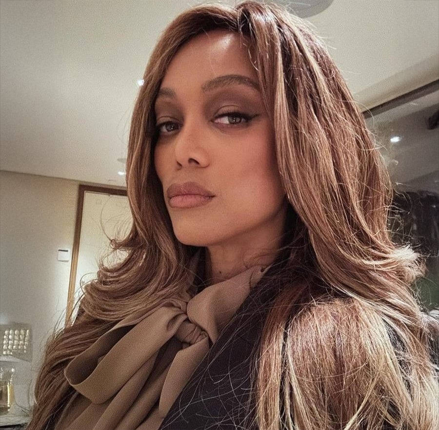 A woman with long, wavy brown hair and a neutral expression poses indoors, wearing a tan blouse with a bow at the neck.
