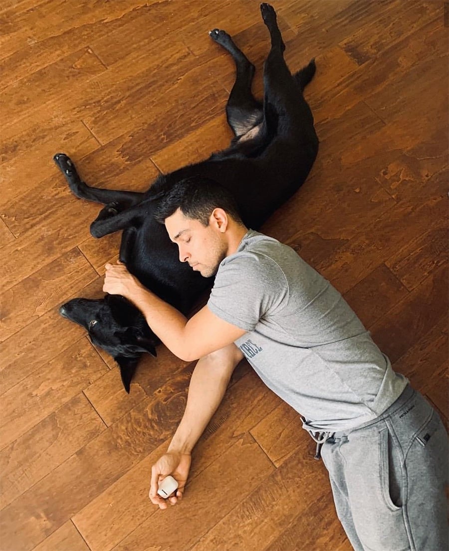 A man in a gray shirt and sweatpants lies on a wooden floor, hugging a large black dog who is also lying on the floor. The man’s eyes are closed, and both appear relaxed and comfortable.