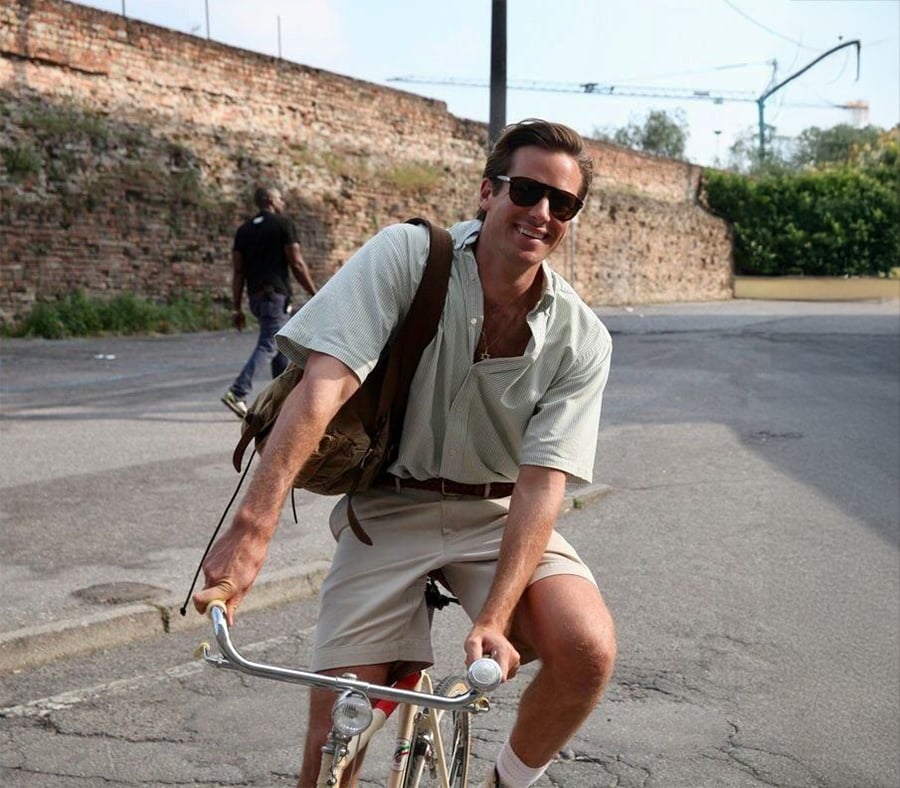 A smiling man wearing sunglasses, a light shirt, and shorts rides a bicycle on a sunny street with a brick wall in the background.