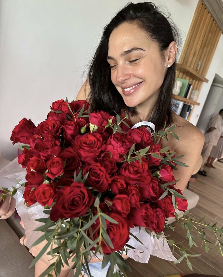 A woman with dark hair smiles while holding a large bouquet of red roses with greenery. She is indoors, and shelves and another person are visible in the background.