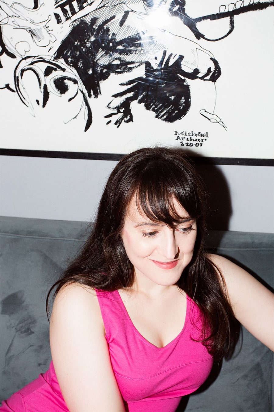 A woman with long dark hair wearing a bright pink top sits on a gray couch, smiling slightly with her eyes closed. Behind her is a black and white art poster on the wall.