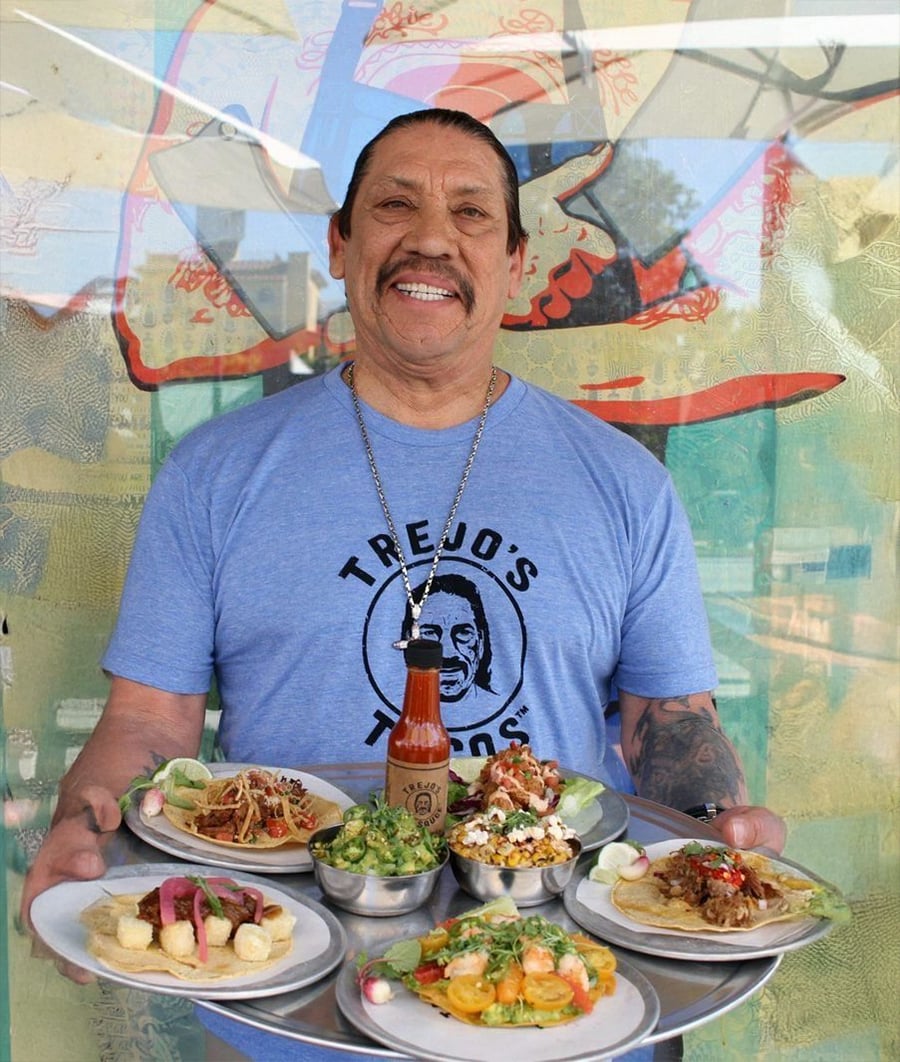 A smiling man in a "Trejo's Tacos" shirt holds a tray with tacos, guacamole, and hot sauce, standing in front of a colorful mural.