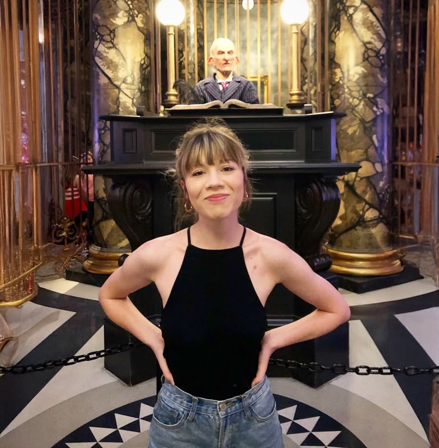 A smiling woman in a black sleeveless top and jeans stands in front of a chained-off area with a goblin banker figure at a desk, inspired by a fantasy movie set.