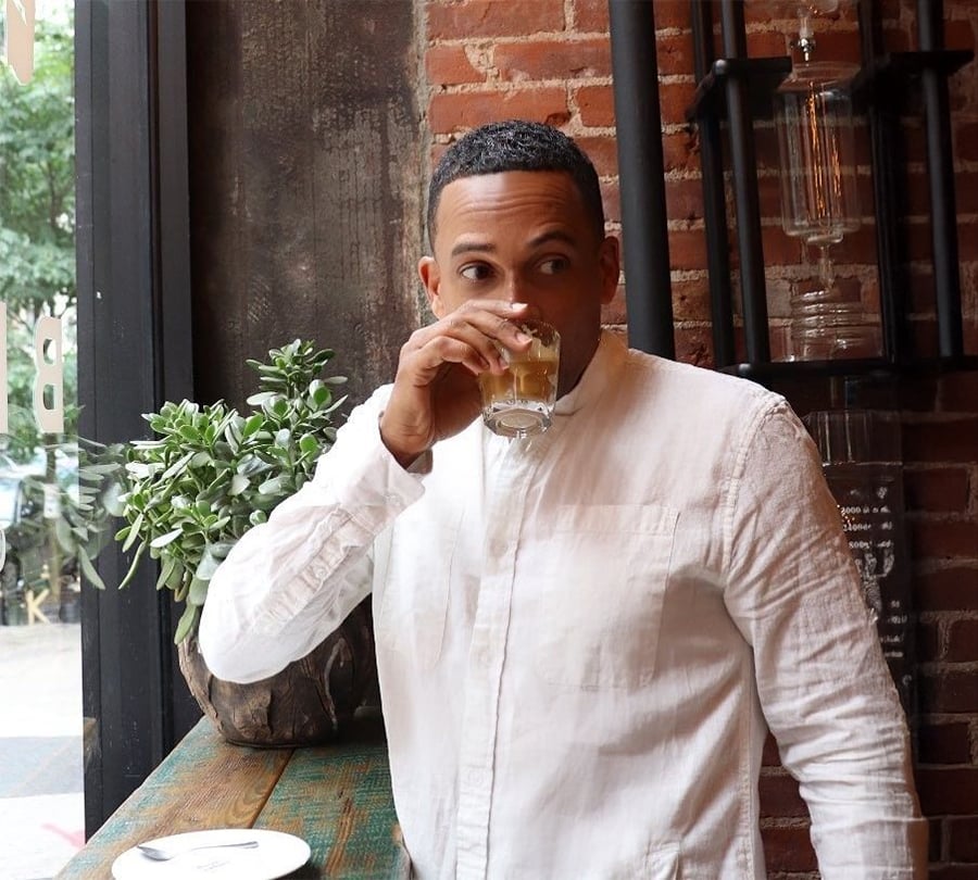 A person in a white shirt drinks from a glass while standing by a window in a rustic cafe with brick walls and green plants.