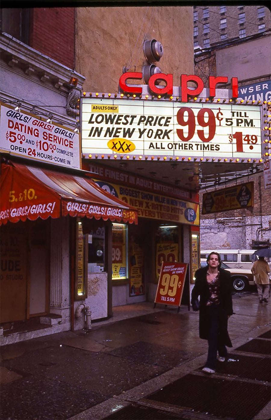 A neon-lit Capri theater advertises adult movies for 99 cents, with signs for a dating service and "GIRLS! GIRLS! GIRLS!" A person walks past on a rainy city street, and old cars are parked nearby.