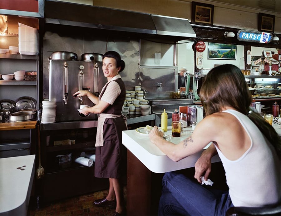 A waitress in uniform smiles while filling cups behind a diner counter. A man with long hair and a tank top sits at the counter with condiments, drinks, and food in front of him. Shelves and diner decor fill the background.