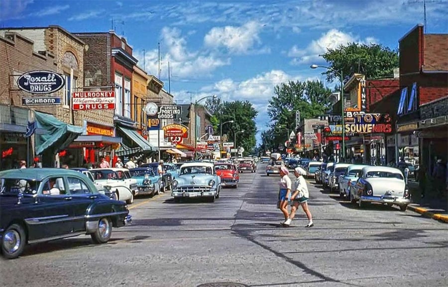 A busy 1950s main street with vintage cars parked along both sides, colorful storefronts and neon signs, and two people crossing the road under a bright, partly cloudy sky.