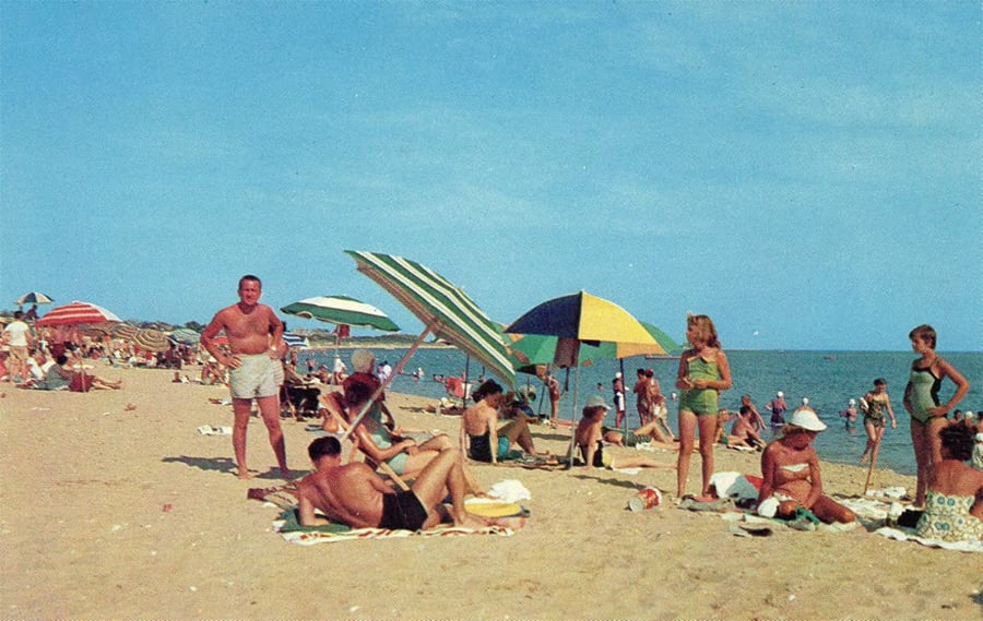 People relax on a sandy beach under colorful umbrellas, with some sunbathing, sitting, or standing. The sky is clear and blue, suggesting a warm, sunny day by the water.