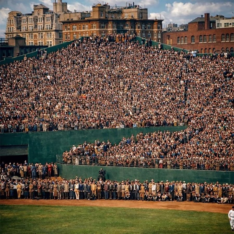 Thousands of spectators fill the stands and line the outfield wall at a baseball stadium, with city buildings visible in the background under a partly cloudy sky.