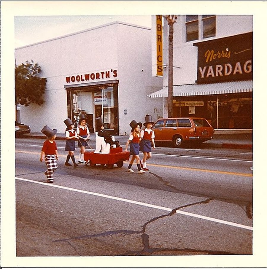 Children in costumes pull a red wagon across a street in front of a Woolworth’s store, with cars and storefronts visible in the background on a sunny day.