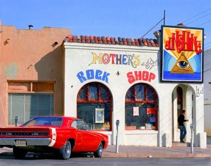 A person enters a shop with colorful signs reading "Mother's Rock Shop." A red vintage car is parked outside and a mural with a pyramid and eye is painted on the building.