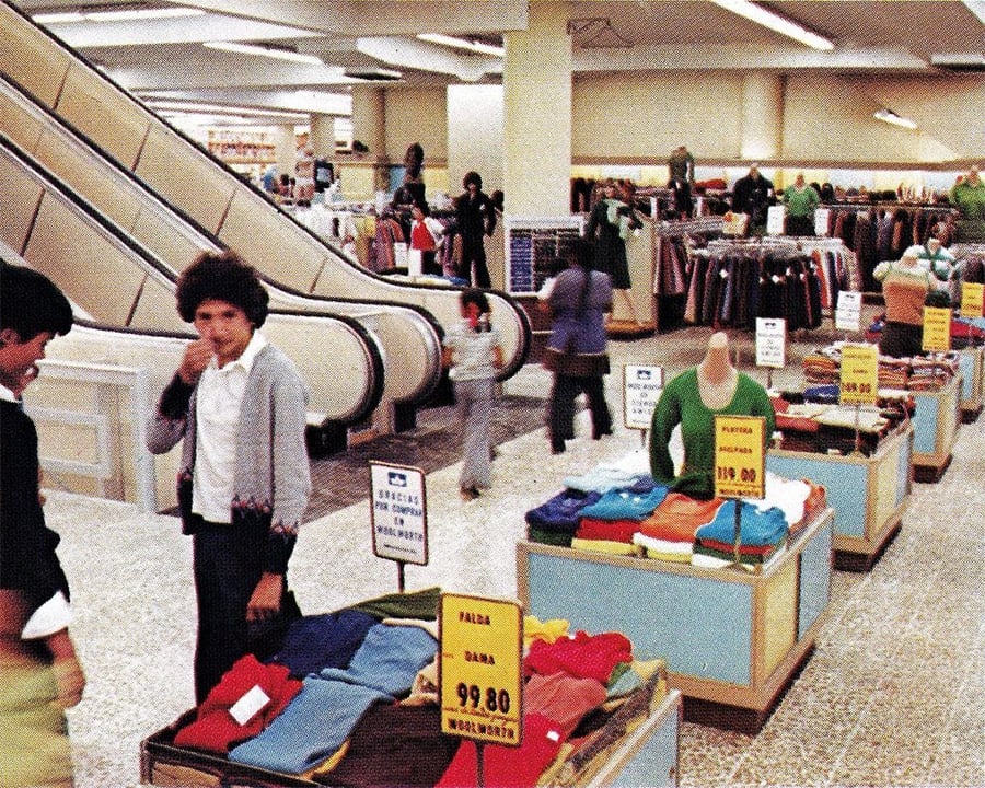 A vintage department store with escalators, clothing racks, and tables displaying folded clothes. Shoppers browse the aisles. Price signs are visible on the tables. The scene looks busy and retro.