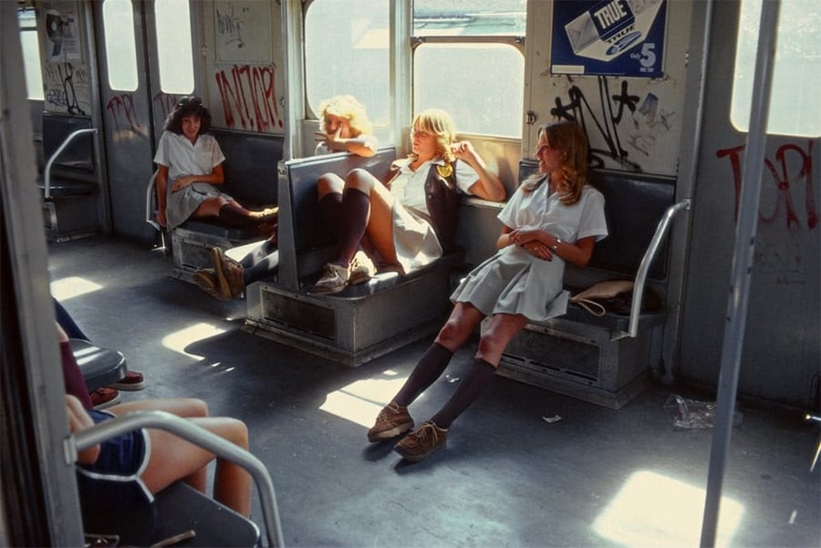 Four girls in school uniforms sit and relax on seats in a graffiti-covered subway car, sunlight streaming through the windows, with two other passengers partially visible on the left.