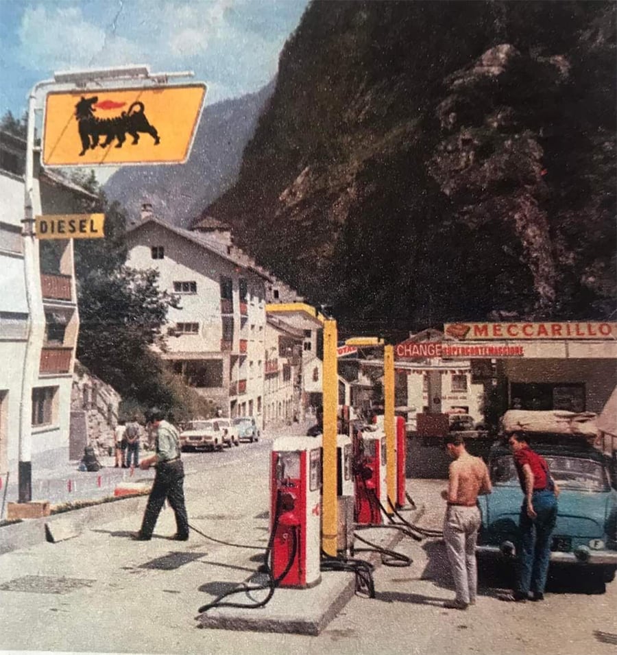 A vintage gas station with yellow pumps, people standing near a blue car, and buildings in the background, set in a mountainous area. Signs read "DIESEL" and "MECCARILLO.