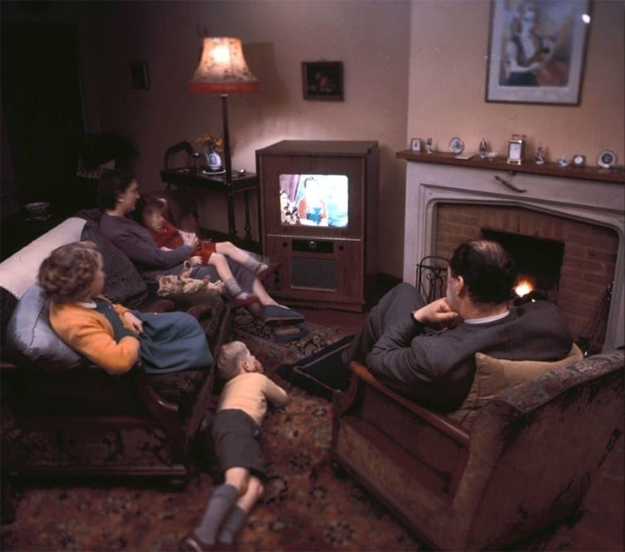 A family of five, including two children, watch TV together in a cozy living room with a lit fireplace, vintage furniture, and warm lighting.