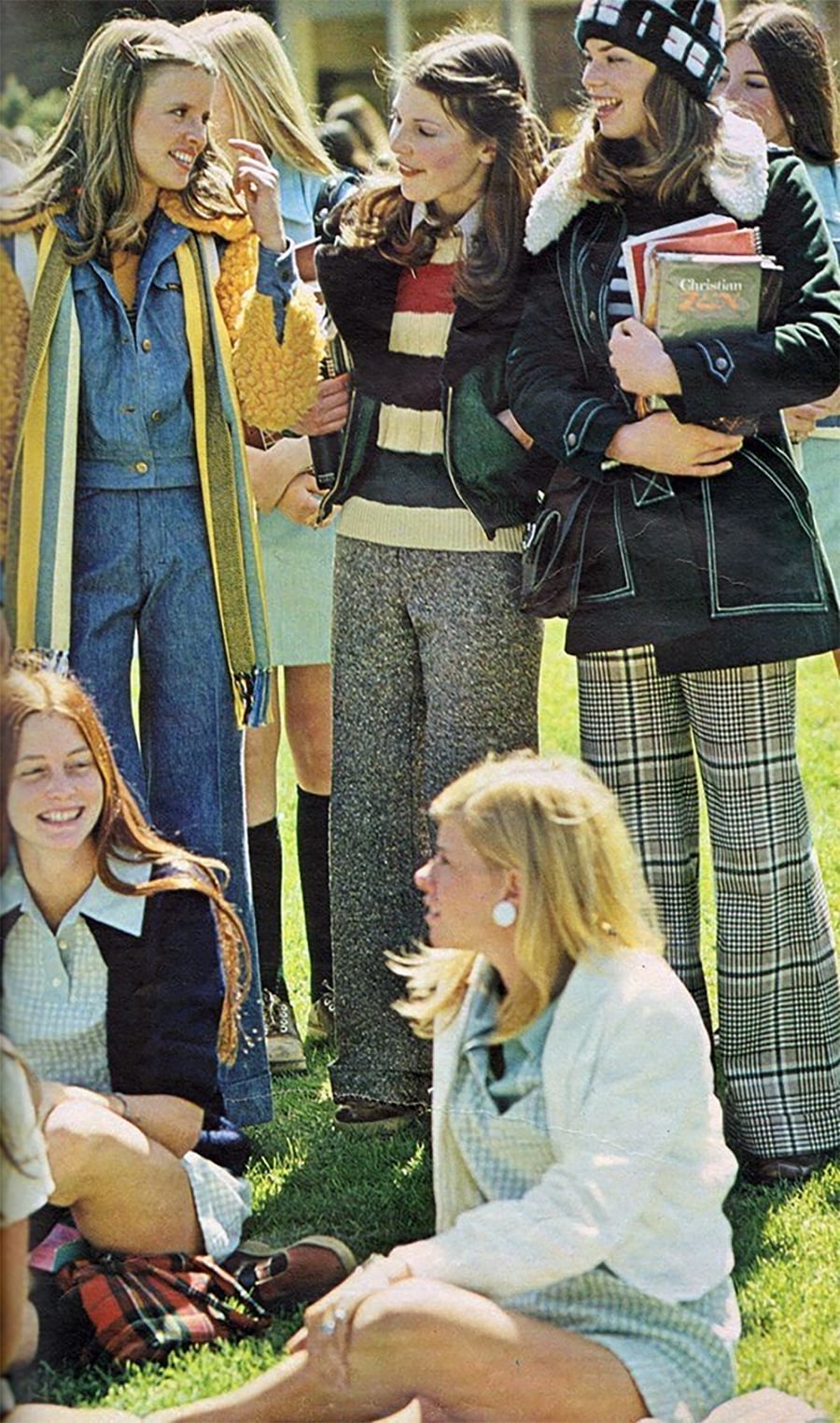 A group of young women in 1970s fashion, including flared pants, sweaters, and jackets, stand and sit on grass, smiling and talking outdoors on a sunny day. Some hold books, suggesting a school setting.