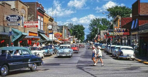 A busy small-town street scene from the 1950s with classic cars parked along storefronts, colorful signs, and two people crossing the street under a bright sky.