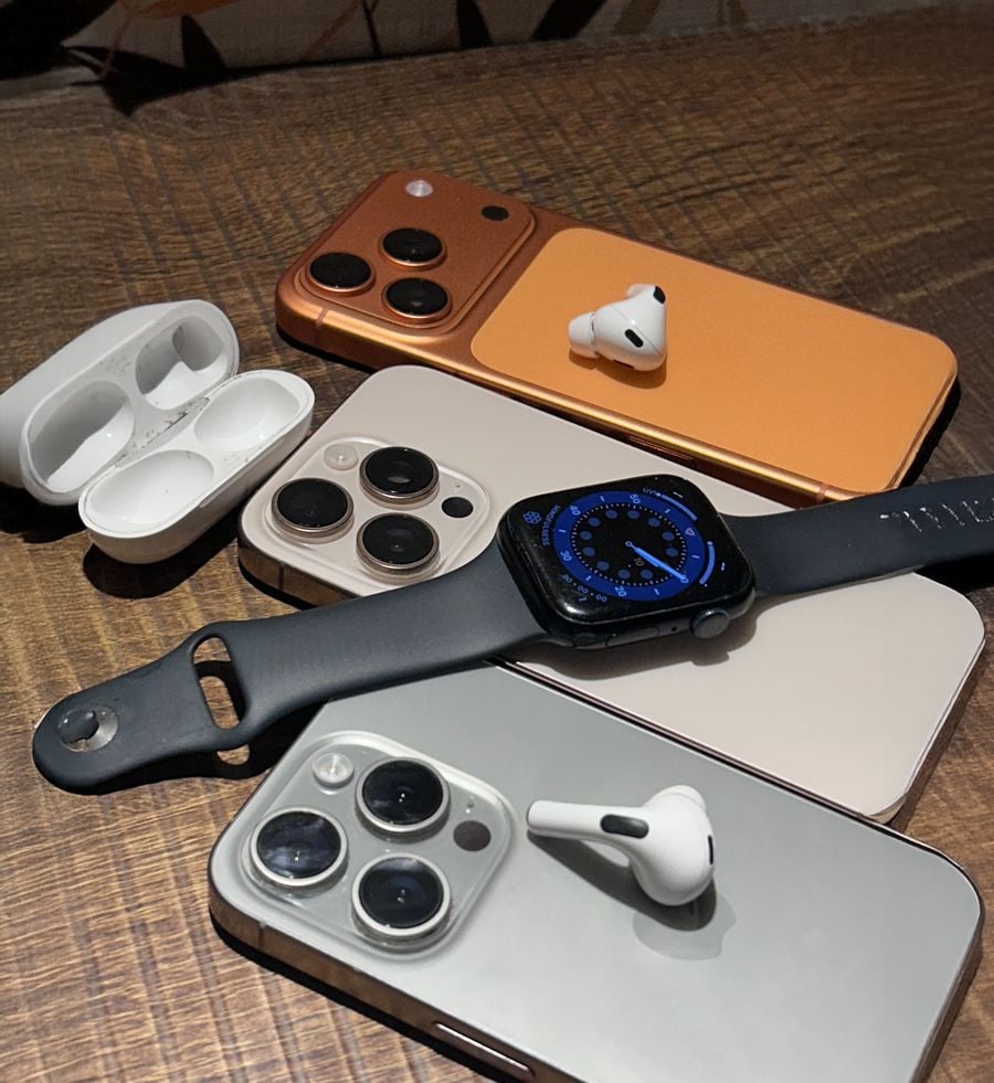 Three iPhones, two AirPods earbuds, an open AirPods case, and an Apple Watch with a black band are displayed on a wooden surface.