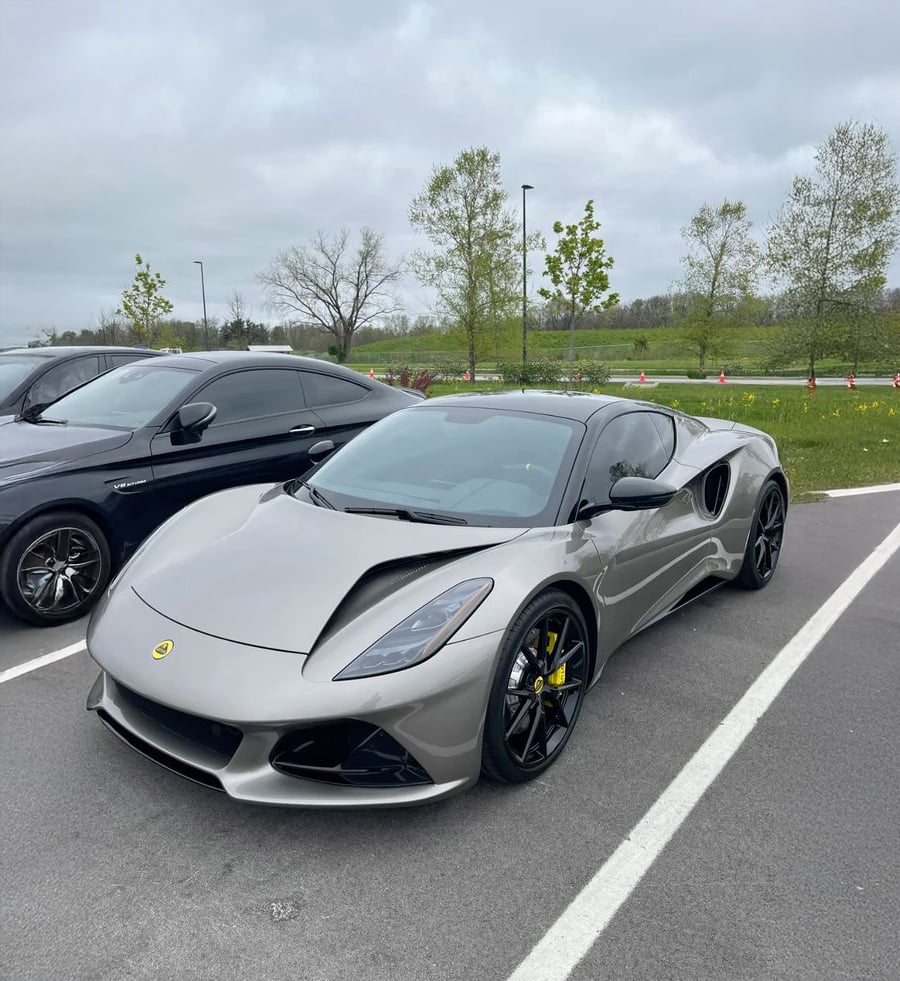 A silver Lotus sports car with black wheels is parked in a lot next to a black car, with green grass, trees, and an overcast sky in the background.
