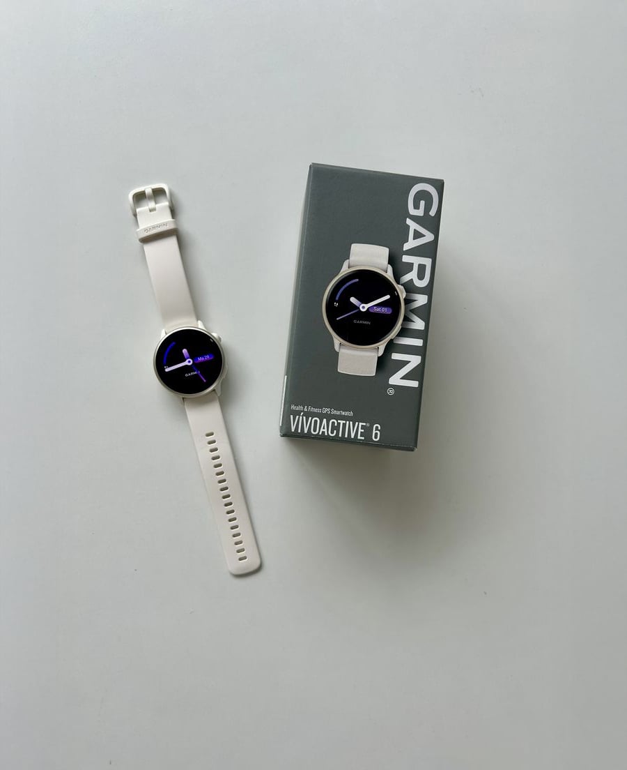 A white Garmin Vivoactive 6 smartwatch is displayed next to its box on a plain light surface. The watch has a round face showing the time.
