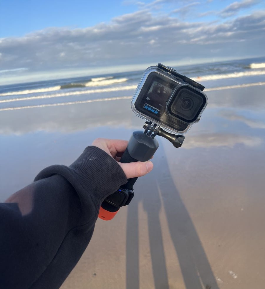 A person holds an action camera on a handle at the beach, pointing it toward the ocean. The sand is wet and reflective, with gentle waves and a cloudy sky in the background.