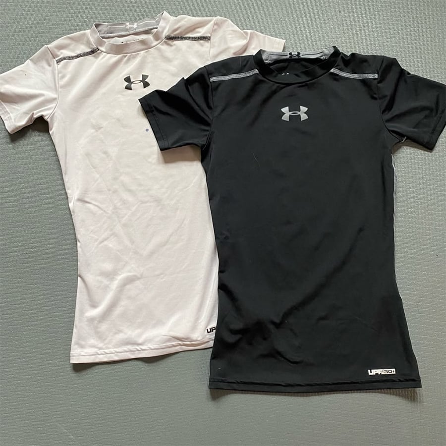 Two Under Armour short-sleeve athletic shirts, one white and one black, lie flat side by side on a textured gray surface. Both shirts display the Under Armour logo on the chest and "UPF 30" at the hem.