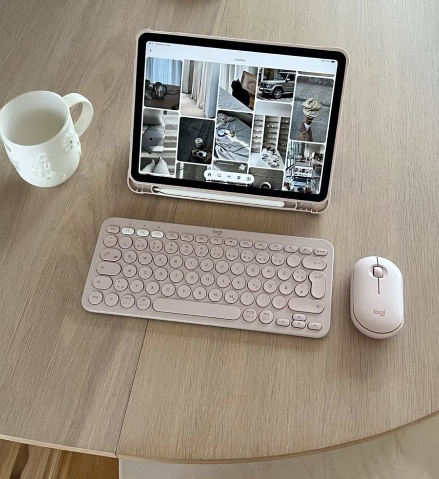 A tablet with a Pinterest screen, a pink wireless keyboard, a pink mouse, and a white mug sit on a round wooden table.