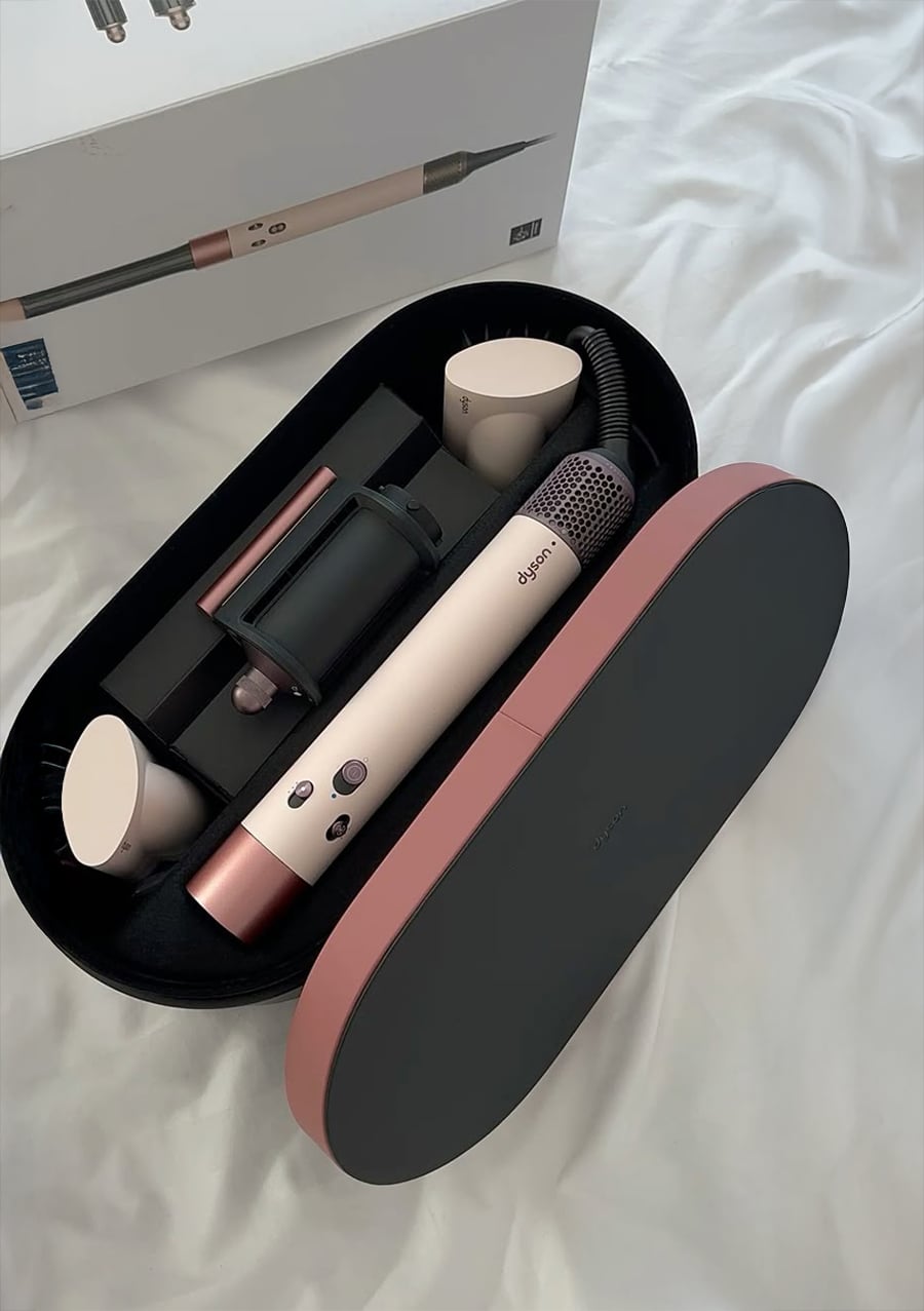 A Dyson Airwrap styler set in a black and pink case with multiple attachments is displayed on a white bed. The product box is visible in the background.