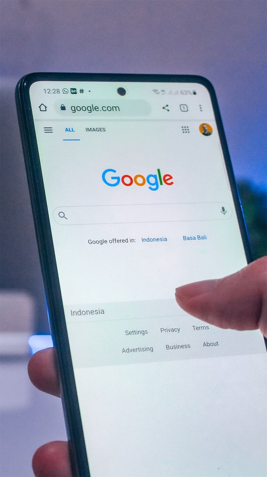 A hand holding a smartphone, displaying the Google homepage set to Indonesia, with the search bar and language options visible on the screen.
