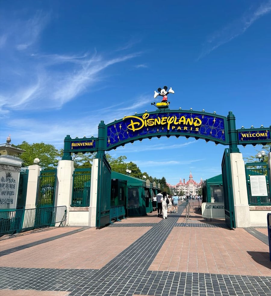 Entrance to Disneyland Paris with a blue archway featuring Mickey Mouse on top, signs reading "Bienvenue" and "Welcome," and people walking toward the park under a sunny sky.