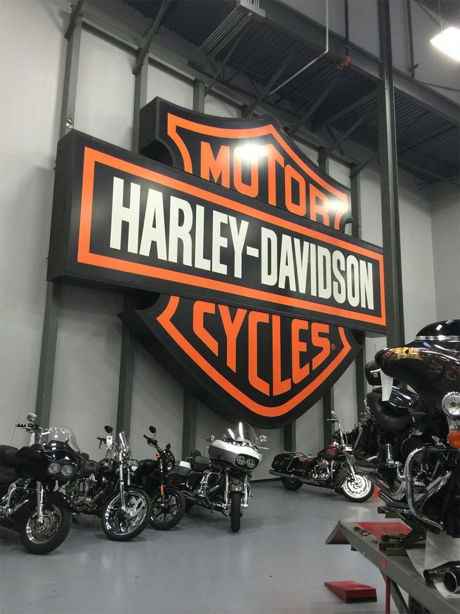 Large Harley-Davidson motor cycles logo sign mounted on a wall above several parked motorcycles inside an indoor showroom or garage.