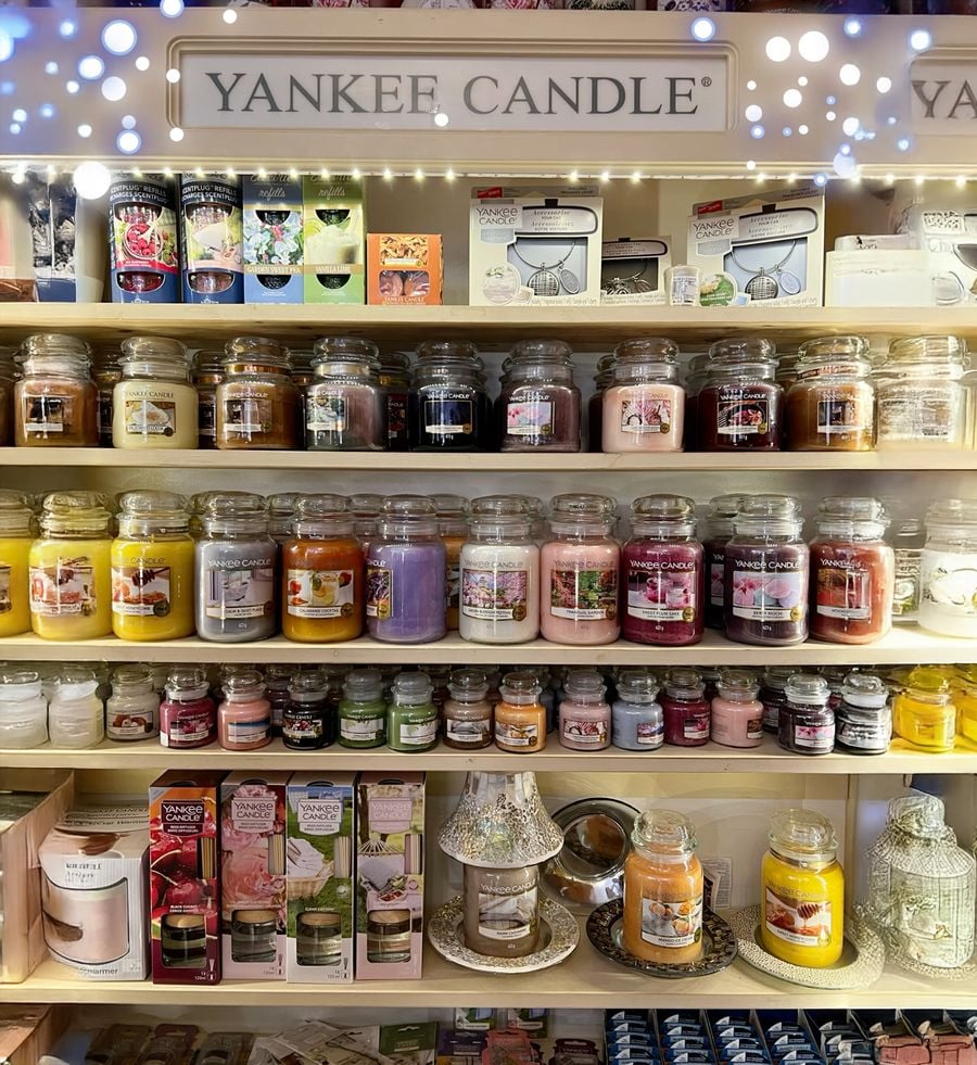 Shelves filled with assorted Yankee Candle jars in various colors and scents, with lit decorative lights above and candle accessories displayed on the bottom shelf.