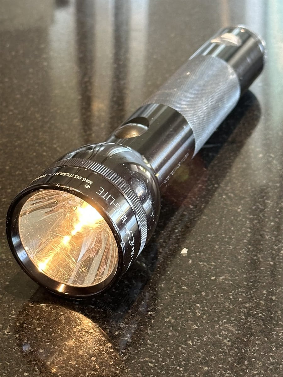 A black Maglite flashlight rests on a dark, reflective surface with its light turned on, casting a bright beam and visible reflections on the table.