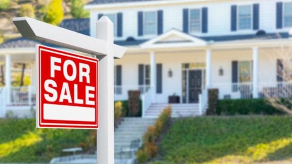 A "For Sale" sign is displayed in front of a large, white two-story house with black shutters, a dark roof, and a front porch, suggesting the home is available for purchase.