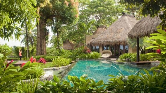 A tropical resort with thatched-roof huts surrounded by lush greenery and colorful flowers, featuring a clear blue swimming pool. A person in a yoga pose stands on a wooden deck in the background.