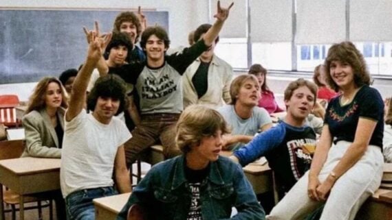 A group of teenagers pose and smile in a classroom, some sitting on desks and chairs. Several make hand gestures, and the mood is lively and cheerful with a blackboard and windows in the background.