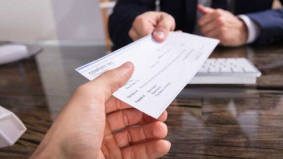 A person hands a check to another person across a wooden desk. A computer keyboard and paperwork are visible on the desk, and both individuals are wearing business attire.