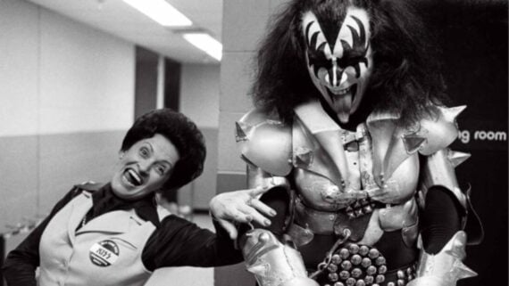 A smiling woman in a uniform playfully holds hands with a person dressed in elaborate spiked armor and black-and-white face paint, posing together in a hallway.