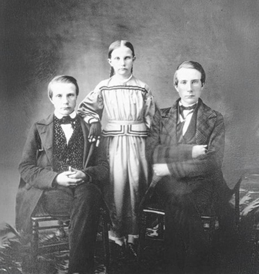 Three children in formal 19th-century attire pose for a black-and-white studio portrait. Two boys sit on chairs, while a girl stands between them. All have serious expressions and light-colored hair.