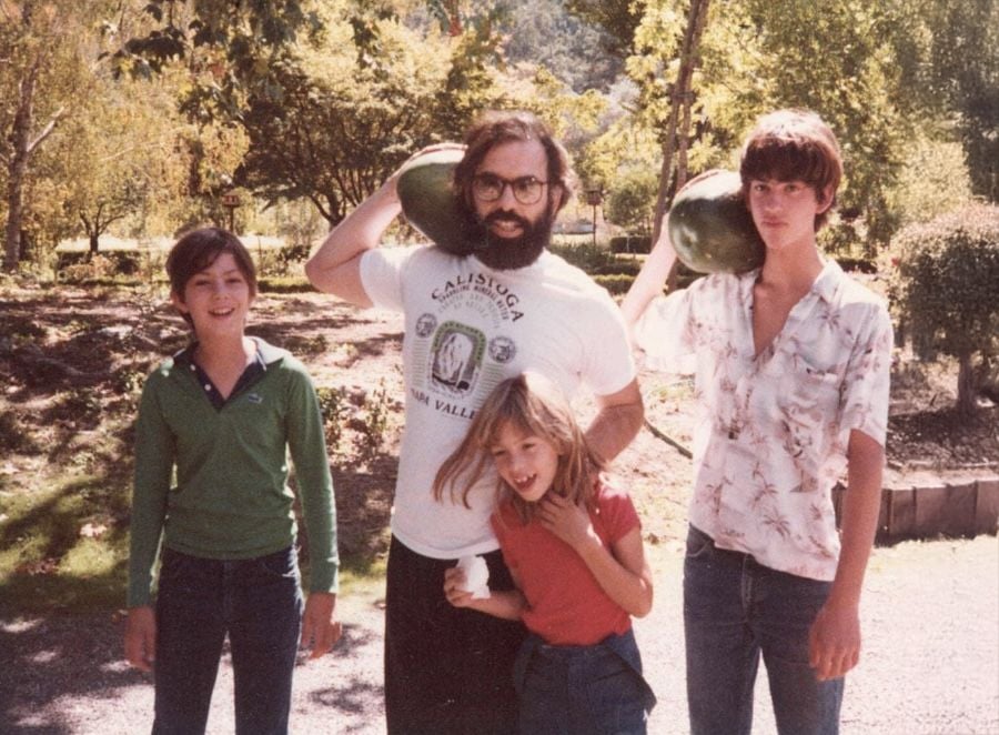 Four people stand outside on a sunny day; the man in the center holds watermelons on his shoulders. Two girls stand on each side, with one hugging a child in front. Trees and greenery are in the background.