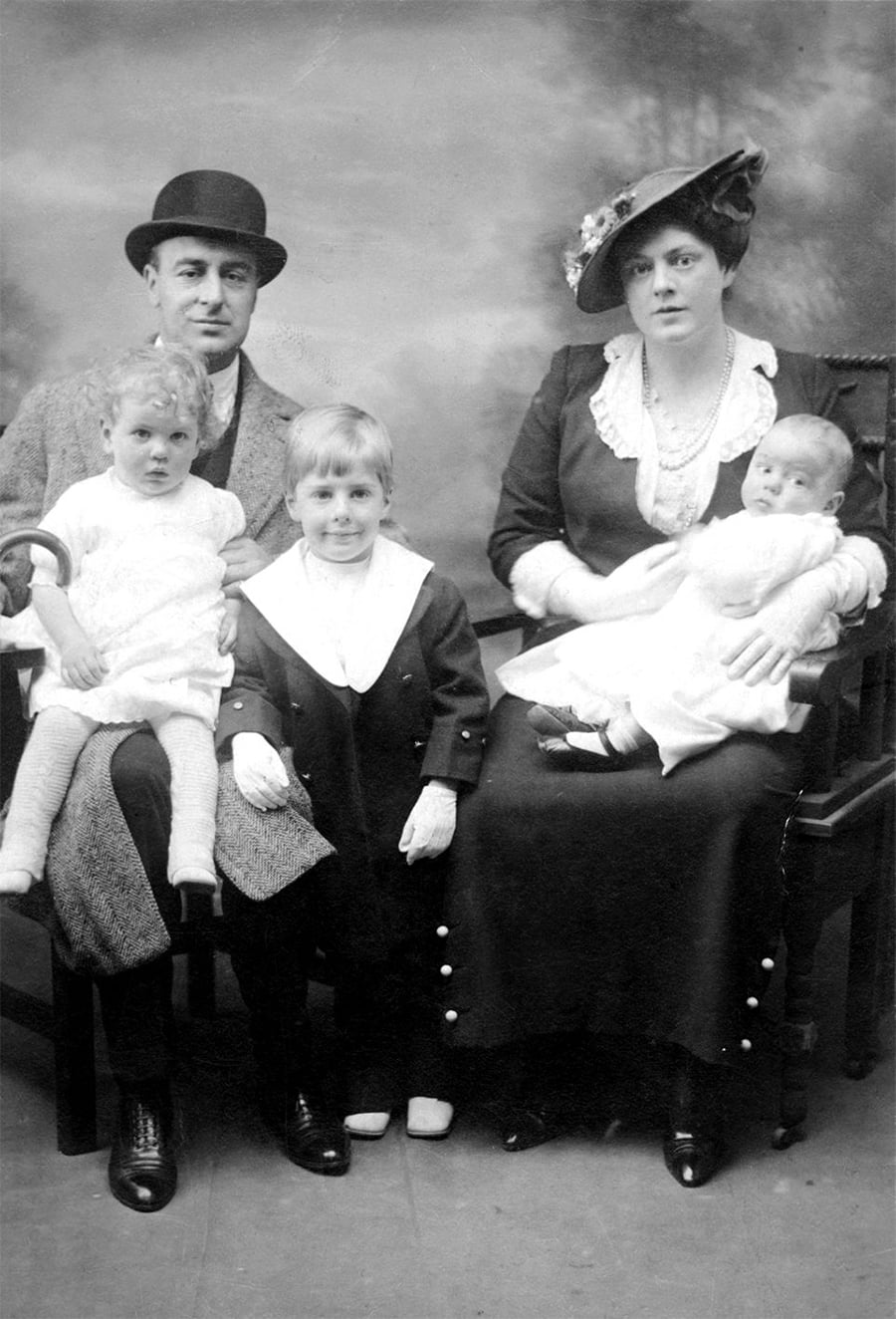 A vintage black-and-white portrait of a family with two adults and three young children. The man and woman are seated, dressed formally, with the children sitting or standing between them, all looking at the camera.