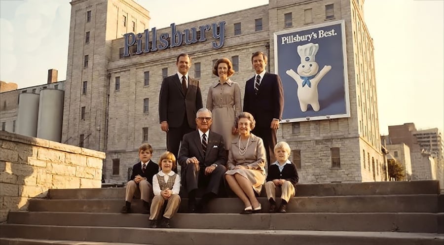 A family poses on stone steps in front of a large building with a “Pillsbury” sign and a billboard featuring the Pillsbury Doughboy. Three adults stand behind four seated people, including three children.