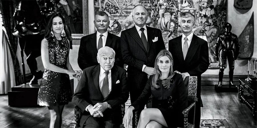 Six well-dressed people pose in an ornate room with artwork, armor, and sculptures. Two are seated in front, while four stand behind them, all looking toward the camera. The photo is in black and white.