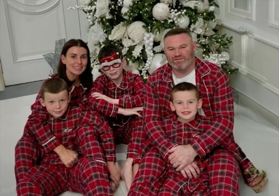 A family of five wearing matching red plaid pajamas sits together by a decorated Christmas tree, smiling at the camera.