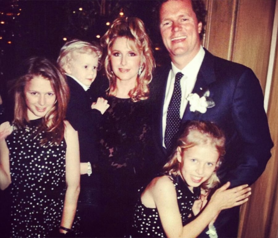 A family of five poses together at a formal event. The man wears a suit with a polka dot tie, while the woman and two girls wear black outfits with white polka dots. The man holds a young child in a suit.